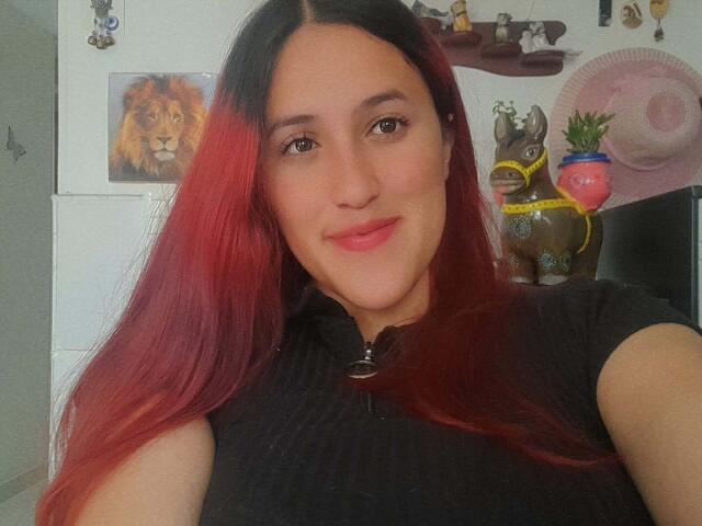 AmbarRossi mit langem, roten und schwarzen Haar, lächelnd, trägt ein schwarzes Hemd und steht vor einer Wand mit einem Löwen. ein Bild, Stofftiere und ein pinker Fächer. Total süß..