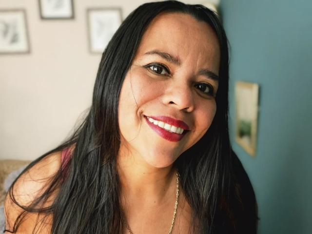 Close-up of VeraCruz with long black hair, smiling with red lipstick, wearing a gold necklace. Blurred background, blue wall on the right, white wall with framed pictures. Fucking hot.