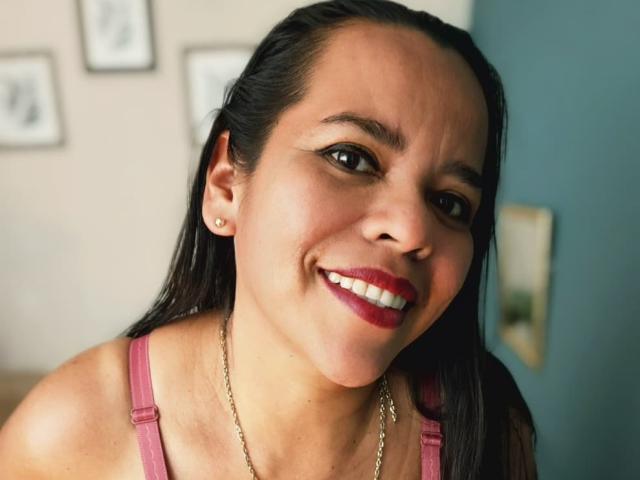 Close-up photograph of VeraCruz with dark straight hair, smiling with red lipstick, wearing a pink tank top and gold necklace. Bright eyes, subtle makeup, blue wall in background. Sexual tension evident.