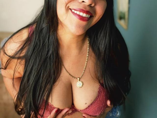 Fotografía de primer plano de VeraCruz, con cabello negro largo, lápiz labial rojo y un collar de oro, sonriendo mientras aprieta su mano. Los senos juntos, en un sujetador rojo de encaje. Escote pronunciado y contornos de los pezones visibles..