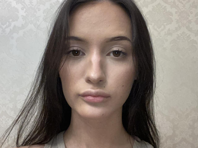 Close-up photograph of MarchelleBerrien with long black hair, neutral expression, light skin, subtle makeup, and small nose piercing. Background is a textured, off-white wall. Focused, slightly intense gaze.