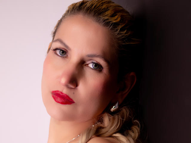 Close-up photograph of GiuliaAddams, fair-skinned with blue eyes, red lipstick, and blonde hair, looking directly at the camera with a sultry expression, heavy shadow on the right side.