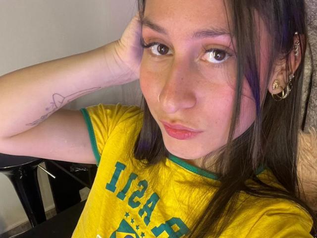 Close-up of MaddyCarina, wearing a yellow "IBA" shirt, head resting on hand, dark brown hair, brown eyes, slightly flushed skin, small hoop earrings, tattoo on upper arm, sexy, sultry expression.