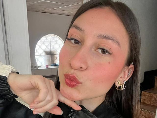 Close-up photo of MaddyCarina with pale skin, dark brown hair, and red lipstick. She's pouting, touching her cheek with her right hand. Wearing a black top, gold hoop earrings. Bright background.