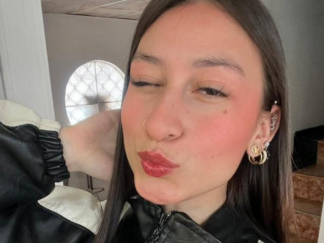 Close-up selfie of MaddyCarina, winking with puckered red lips, black nose ring, gold hoop earrings, long straight brown hair, black jacket, bright light in background, soft natural light on face.