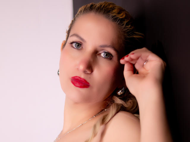 Close-up of GiuliaAddams with striking red lipstick, blue eyes, and fair skin. She's touching her face with one hand, wearing a pearl necklace. Dark background contrasts her light complexion. Sexual tension evident.