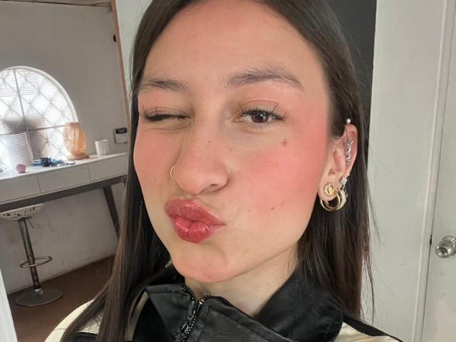 Close-up selfie of MaddyCarina with fair skin, winking, and puckering lips. She has dark brown hair, a nose ring, and gold earrings. Wearing a black top, cocky expression. Bright room background.