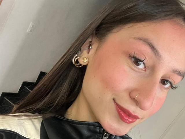 Close-up selfie of MaddyCarina, light-skinned with brown hair, wearing a black jacket, gold hoop earrings, and red lipstick, smiling subtly, right eye slightly closed, slight blush on cheeks.