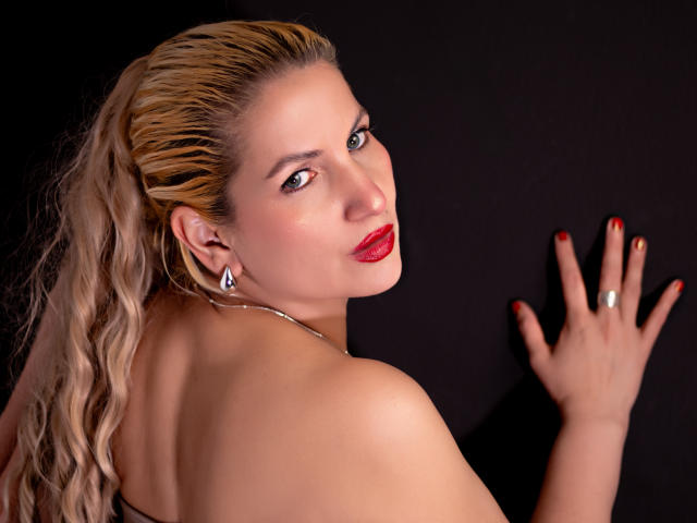Foto van GiuliaAddams, met de rug naar de camera, die over haar schouder kijkt, rode lippen, glad achterover gekamd blond haar, zilveren oorbellen. rode nagellak, linker hand tegen donkere achtergrond, sexy, intense blik, verleidelijke uitdrukking.