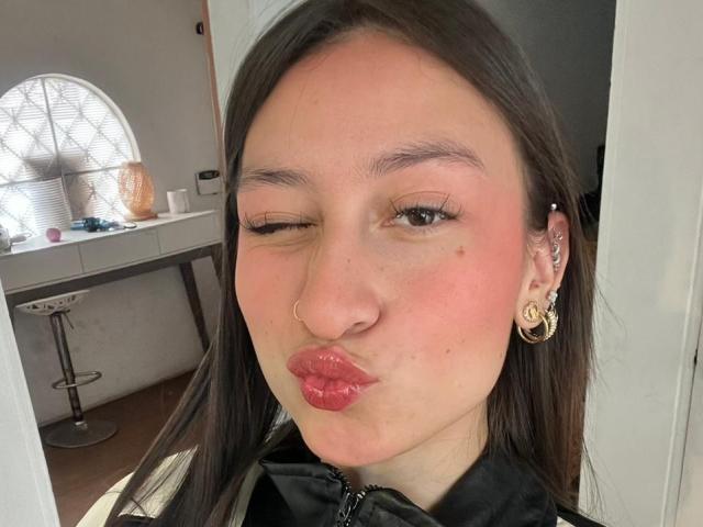 Close-up selfie of MaddyCarina with one eye closed, puckering lips for a kiss. She has long dark hair, a nose ring, gold hoop earrings, and a black top. Background shows a desk and window.
