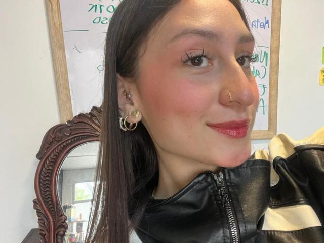 Close-up van MaddyCarina, een jonge vrouw met lang donker haar, rode lippenstift en gouden oorringen, die een zwarte leren jas. Ze heeft een neusring en een zelfverzekerde glimlach..