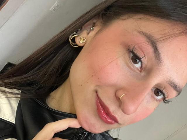 Close-up photo of MaddyCarina, smiling with red lipstick, brown eyes, and a gold nose ring. She has long dark hair, wears gold earrings, and touches her chin. Sexual tension from direct gaze.