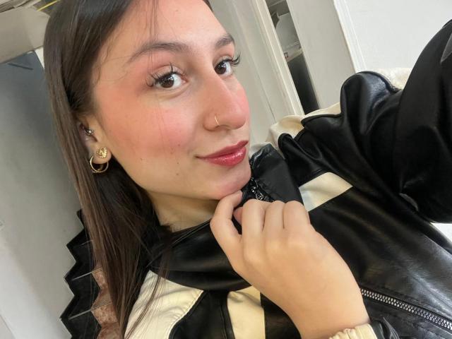 Close-up selfie of MaddyCarina with light skin, brown hair, and red lipstick. She's wearing a black leather jacket with a beige stripe, holding her collar with one hand. Flirtatious smile.