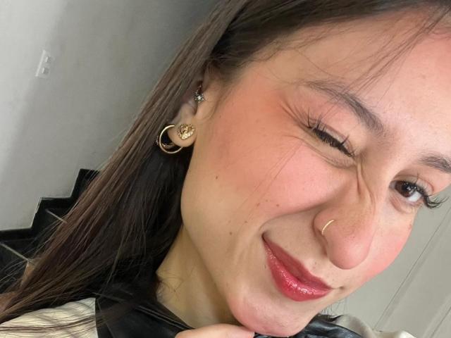 Close-up of MaddyCarina with winking right eye, red lipstick, and a nose ring. She has long brown hair, gold hoop earrings, and a black shirt. Fucking cute, slightly playful expression.