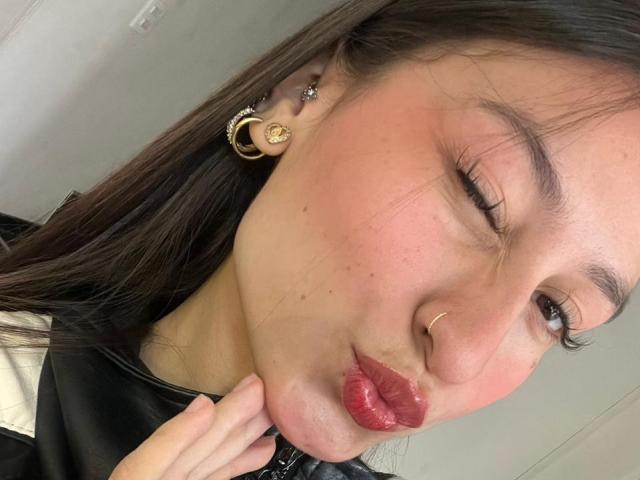 Primer plano de MaddyCarina, una mujer asiática con cabello largo y negro, un ojo cerrado, labios rojos fruncidos, aretes de aro dorados, nariz. pinchando, tocando su cuello, piel con pecas, intensa tensión sexual.