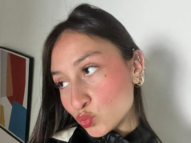 Close-up of MaddyCarina, a young woman with long black hair, pouty red lips, and flushed cheeks. She's wearing gold earrings and a black top, looking sideways with a flirtatious expression.
