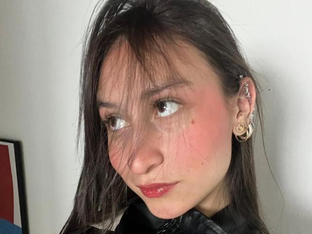 Fotografia ravvicinata di MaddyCarina, una giovane donna con lunghi capelli neri, labbra rosse e guance arrossate, che guarda leggermente di lato. un lato con un'espressione sottile e seducente. Indossare orecchini dorati.