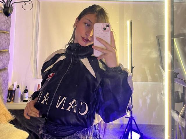 MaddyCarina macht ein Selfie vor einem Spiegel, trägt eine schwarze Jacke mit weißen Buchstaben und hält ein Handy mit einer rosa Hülle. Spiegelbild, das helles Licht und Toilettenartikel auf einem Regal zeigt..