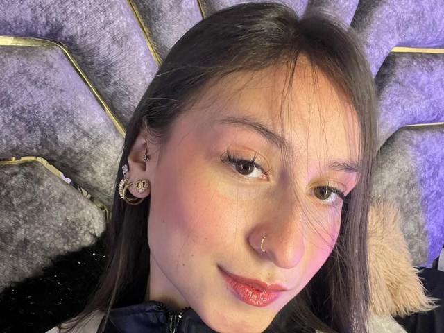 Close-up photo of MaddyCarina, fair-skinned woman with black hair, wearing a black jacket, gold hoop earrings, and a nose ring. Her lips are glossed with red lipstick, smiling slightly. Fucking sexy.