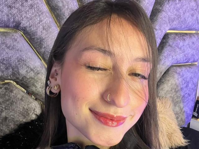 Close-up of MaddyCarina, eyes closed, smiling, with a nose ring and earrings. She has straight brown hair, light skin, and red lipstick. Wearing a black coat with a fur collar.