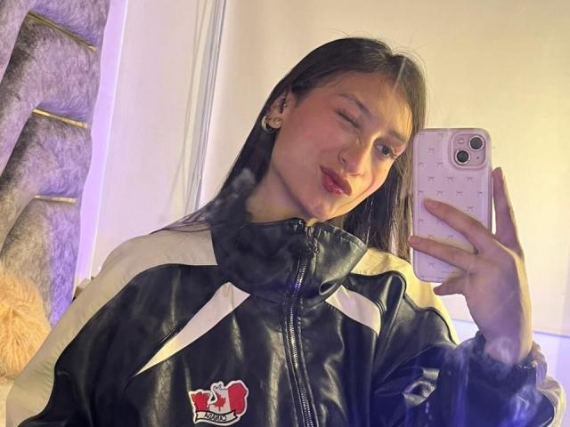 MaddyCarina, a young woman with long black hair, takes a selfie in a mirror. She's wearing a black and white jacket with a red logo, holding a white phone. She's winking and has red lipstick. Her hand is on her cock.
