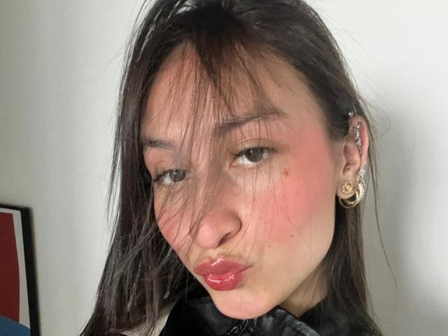 Close-up of MaddyCarina, Asian woman with long black hair, pink cheeks, and glossy red lips. She wears gold earrings, has a slightly flushed face, and a sultry expression. Bright white background.