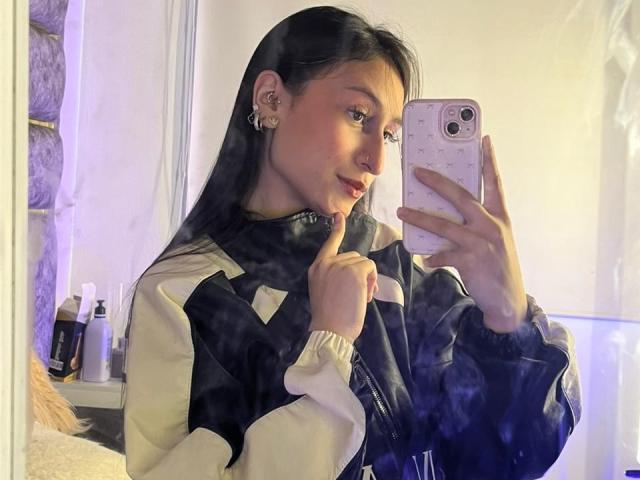 MaddyCarina with long black hair, holding a phone in her right hand, thumb up to her lips, wearing a black and white jacket, taking a selfie, light blue background.
