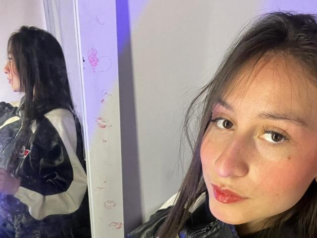 Photograph of MaddyCarina, two views. Left: side profile, black jacket, long dark hair, white sleeves, shadowed face. Right: close-up, face, brown eyes, red lips, slight smirk, light skin. Sexual tension, casual pose.