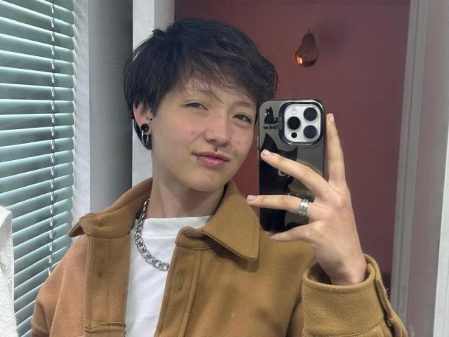 LissyLuxe, a butch-looking person with short black hair, takes a mirror selfie with an iPhone. Wearing a brown coat over a white shirt, they smirk slightly. Bright light from blinds.