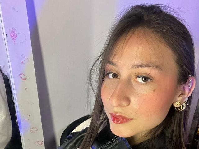 Close-up selfie of MaddyCarina, an Asian woman with long black hair, red lipstick, and gold earrings, looking directly at the camera with a subtle, suggestive smile. Bright lighting, minimal background.