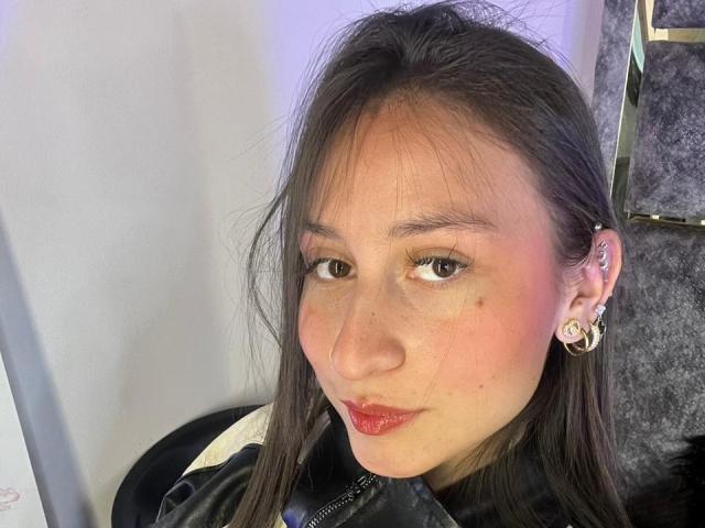 Close-up of MaddyCarina, an Asian woman with long black hair, freckles, and red lipstick. She's wearing a black jacket, hoop earrings, and looking slightly to the side with a seductive expression.