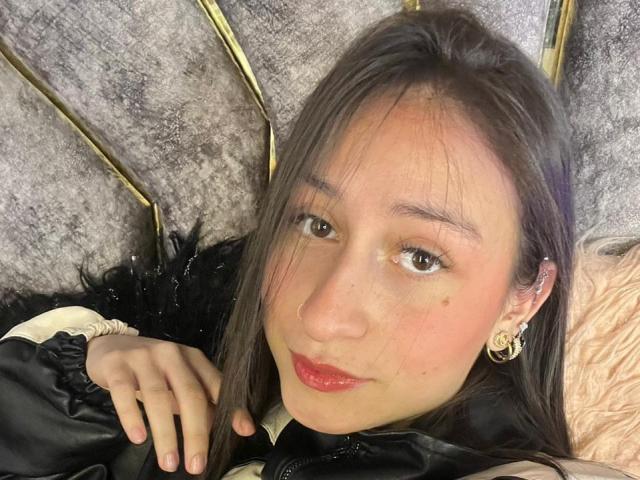 Close-up of MaddyCarina, Asian woman with long black hair, red lipstick, and gold earrings, lying on a grey, textured surface. Her hand rests on a black garment, looking directly at the camera.