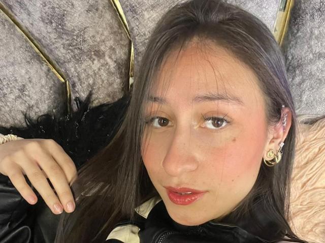 Close-up van MaddyCarina, een Aziatische vrouw met lang zwart haar, rode lippenstift en gouden oorbellen, liggend op een grijze, pluizige oppervlak. Haar hand rust op haar zwarte jas, en ze kijkt recht in de camera. Haar uitdrukking is lichtelijk verleidelijk..