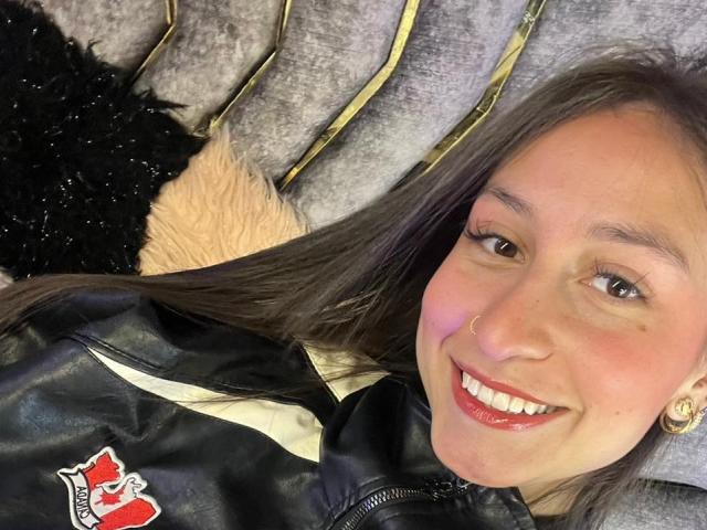 Close-up photo of MaddyCarina with long brown hair, smiling, wearing a black leather jacket with a red patch, lying on a grey and black textured surface, showing sexual tension.