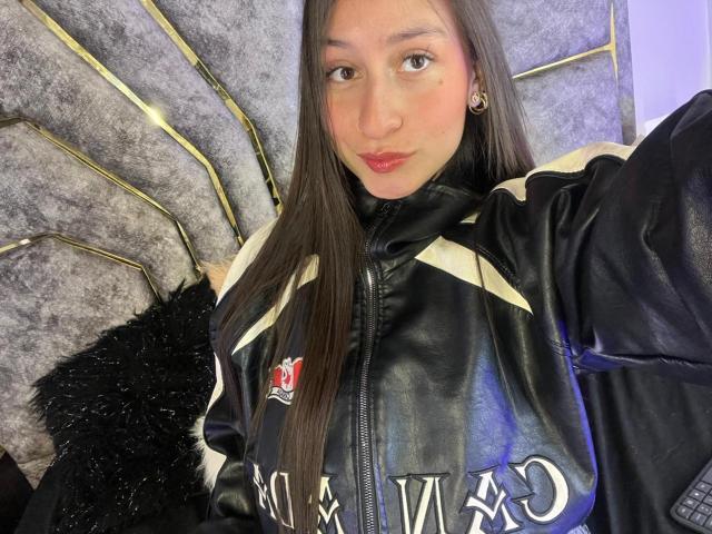 Close-up of MaddyCarina, wearing a black leather jacket with white and red logos, against a gray, patterned wall. She has long black hair, pink lips, and a neutral expression. Fucking sexy.