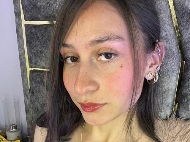 Close-up of MaddyCarina, a young woman with long black hair, fair skin, and red lipstick, wearing a nose ring and large earrings. Her expression is neutral, with slight pink blush on her cheeks. Her breasts are partially visible.