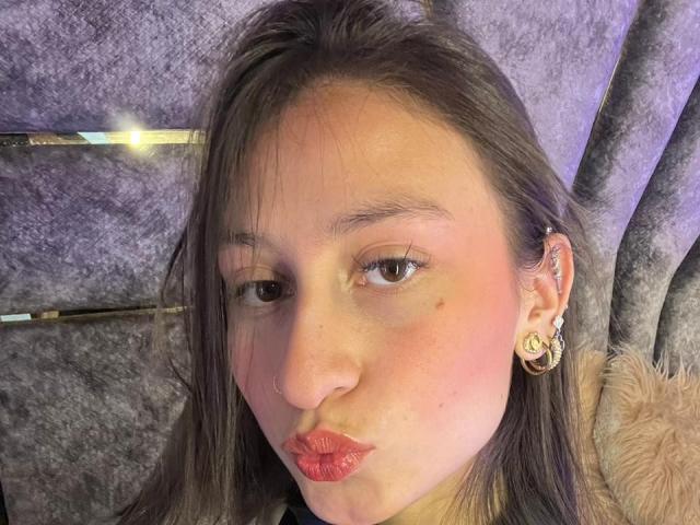 Close-up van MaddyCarina met glanzende rode lippen, donker steil haar en gouden oorbellen, gekleed in een bontgevoerd jasje, kijkend. Zijwaarts, met een verleidelijke uitdrukking, zachte paarse achtergrond. Heel erg aantrekkelijk..