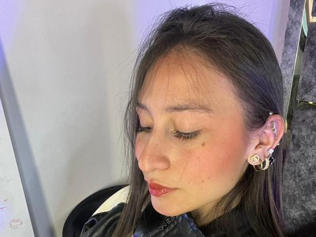 Close-up of MaddyCarina, an Asian woman with long black hair, wearing a black top, red lipstick, and floral earrings. Her eyes are closed, showing freckles and light makeup. Her expression is calm.