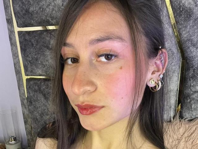 Close-up of MaddyCarina, light-skinned Asian woman with straight black hair, red lipstick, and round earrings, looking directly at the camera with a slightly sexy, serious expression. Bright lighting highlights her face.