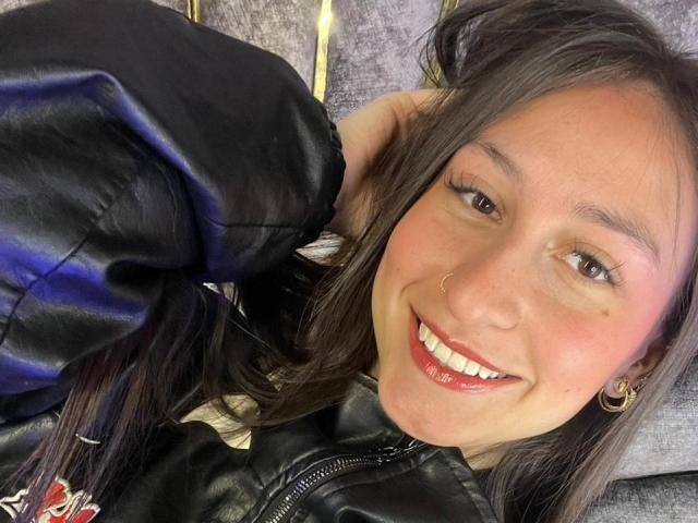 Nahaufnahme von MaddyCarina mit einem strahlenden Lächeln, rotem Lippenstift und dunkelbraunen Augen. Sie trägt eine schwarze Jacke, goldene Ohrringe im Stecker-Design und berührt ihr Haar. Verdammt süß, sexy Aussehen..