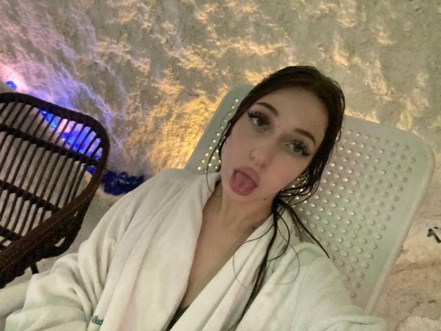 SquirtOfAngel, avec ses cheveux noirs et humides, sa langue tirée, portant une robe blanche, assis sur une chaise percée, mur texturé. fond, lumières bleues et oranges, sexy, provocateur, légèrement flou, selfie décontracté.