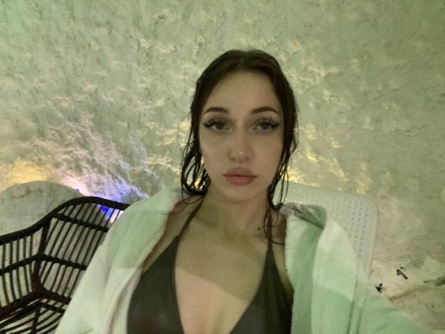 SquirtOfAngel hat nasses Haar, viel Make-up und einen schwarzen Bikini-Oberteil, und blickt direkt in die Kamera mit ernstem Gesicht. Ausdruck. Sie trägt eine weiße Jacke über ihren Schultern. Schwach beleuchteter, strukturierter Hintergrund..