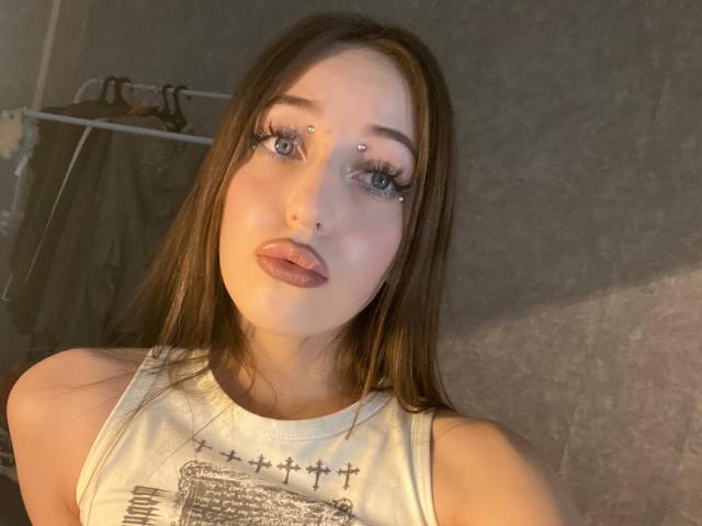 Autoportrait en gros plan de SquirtOfAngel, une jeune femme avec de longs cheveux bruns, des yeux bleus et des piercings faciaux. Elle porte un débardeur blanc et regarde directement l'appareil photo avec une expression séduisante..