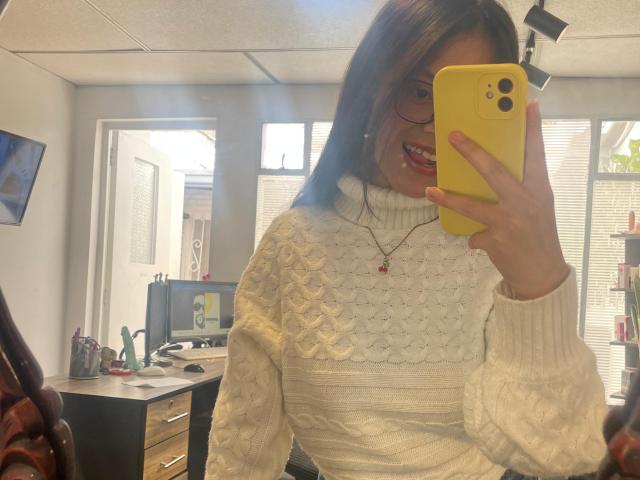AnittaRouses maakt een selfie met een gele iPhone, en draagt een witte, gebreide trui met een structuur. Ze heeft bruin haar dat tot op haar schouders reikt, rode lippen en een rode ketting. Bureau op de achtergrond. Ongelooflijk aantrekkelijk..