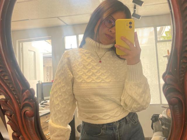 AnittaRouses, wearing a white cable-knit sweater and high-waisted jeans, takes a mirror selfie with a yellow phone. She has glasses, medium-length brown hair, and a red necklace. Fucking hot.