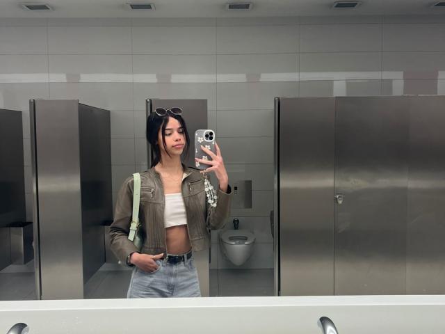 LiaBlom macht ein Selfie in einem modernen Badezimmer mit grauen Fliesen. Sie trägt eine braune Jacke mit Streifen, ein weißes Crop-Top und hoch taillierte Jeans. Schwarzes Haar, Sonnenbrille auf dem Kopf, hält ein Telefon mit schwarzer Hülle..