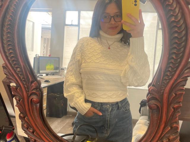AnittaRouses, with shoulder-length brown hair and glasses, takes a mirror selfie. Wearing a white knit sweater and high-waisted jeans, hand in pocket, yellow phone, sexy, confident pose. Ornate mirror frame.
