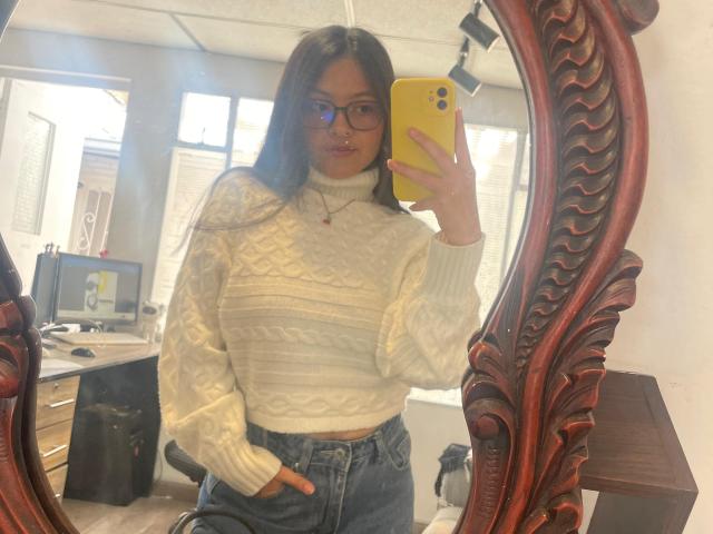AnittaRouses, com cabelos longos e escuros e óculos, tira uma selfie em frente ao espelho, segurando um telefone amarelo. Ela veste um suéter branco de malha e jeans de cintura alta, com uma mão no bolso. Reflete uma sala iluminada..