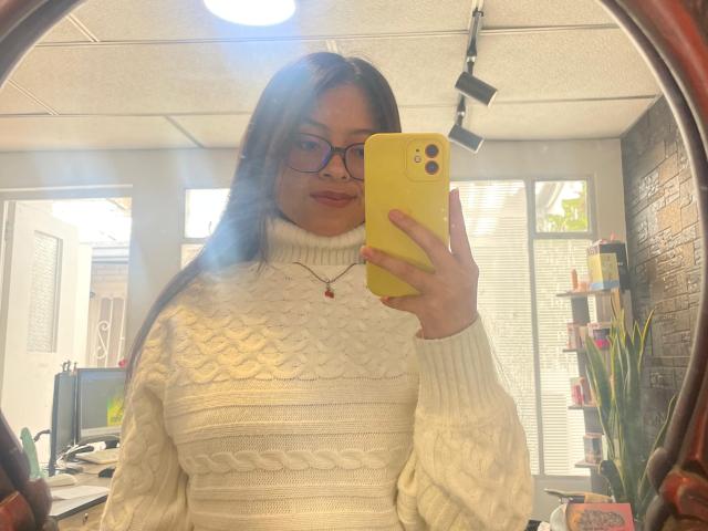 AnittaRouses, wearing a thick white cable-knit sweater, takes a mirror selfie with a yellow phone. She's holding it in front of her chest, flashing a subtle smile. Bright office light.