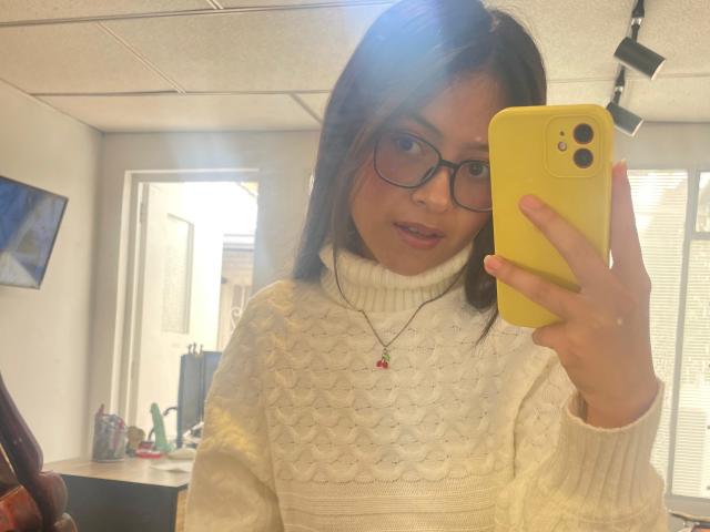 AnittaRouses, wearing a white knitted sweater with a turtleneck, holding a yellow phone, taking a mirror selfie. She has glasses and a pink necklace, with visible sexual tension. Bright office background.